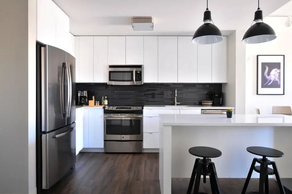 Rental Property-Freshly painted white kitchen, with a black tiled back-splas,sink and fridge,,breakfast bar with black round stools, dark lightshadeswith a image of swan hangingon the wall.