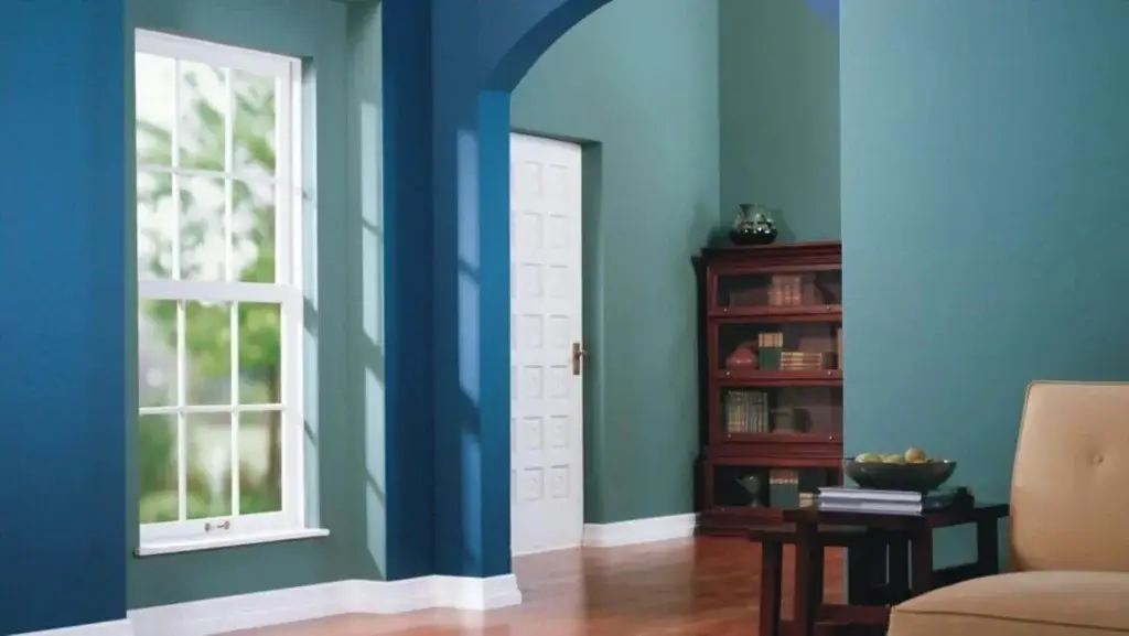 Elegant living room with teal and blue painted walls, featuring a large window, white door, wooden bookshelf, and a beige armchair, showcasing a fresh interior design with vibrant colors